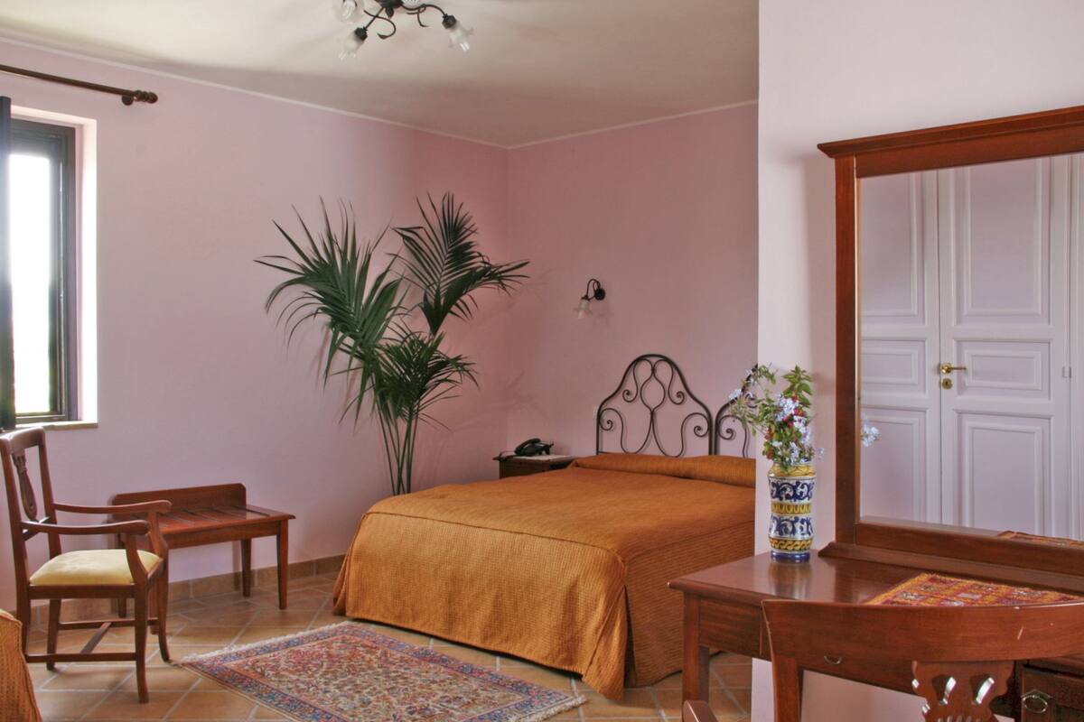 Un dormitorio con cama, una silla, un escritorio, una planta en maceta y una puerta.