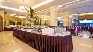 Uma sala de banquetes com uma longa mesa coberta por um toalha castanha, rodeada por cadeiras, e decorada com arranjos florais.