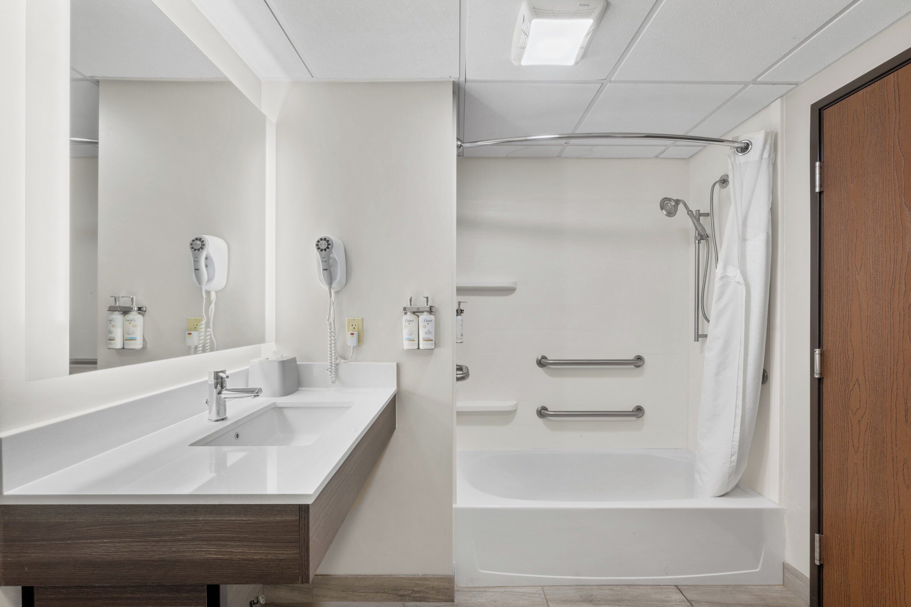 A modern bathroom with a double sink vanity, a bathtub with a shower curtain, and a wooden door.