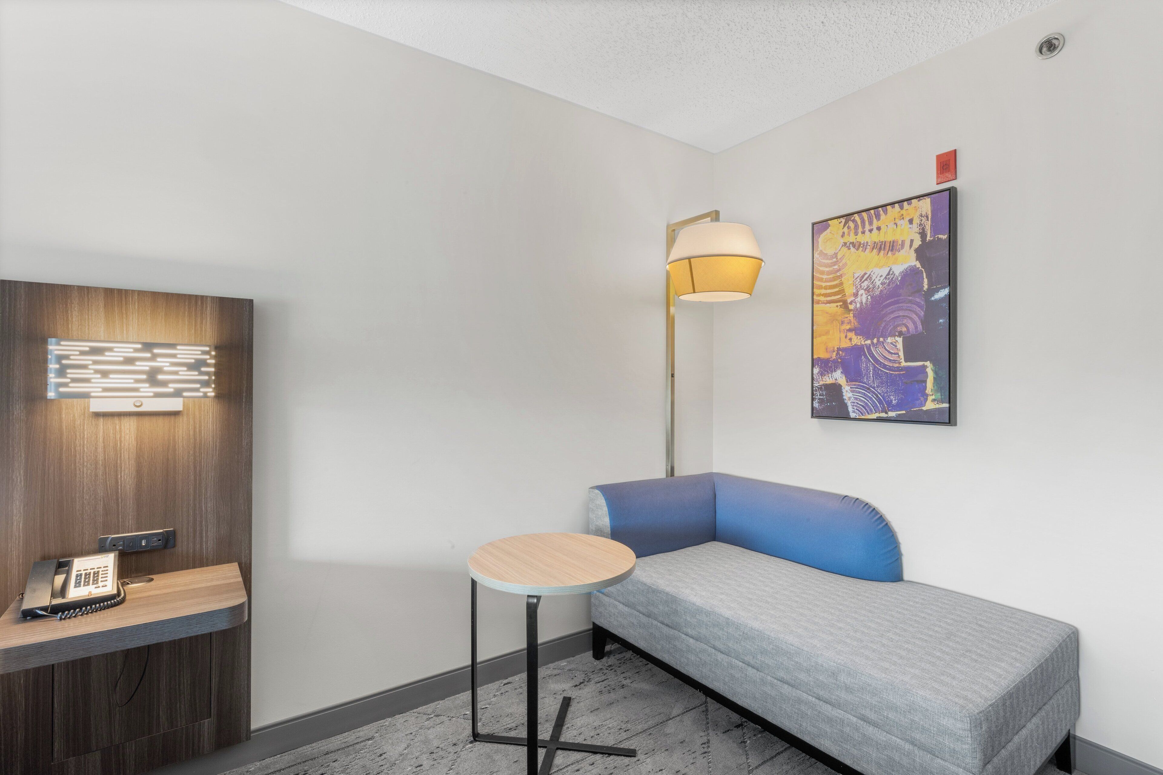 A small, modern room with a grey sofa, a small round table, a lamp, a phone on a wooden shelf, and a colorful abstract painting on the wall.