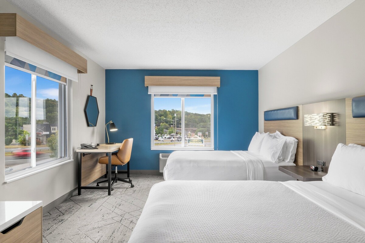 A hotel room with two beds, a desk, a chair, and a large window offering a view of the outside.