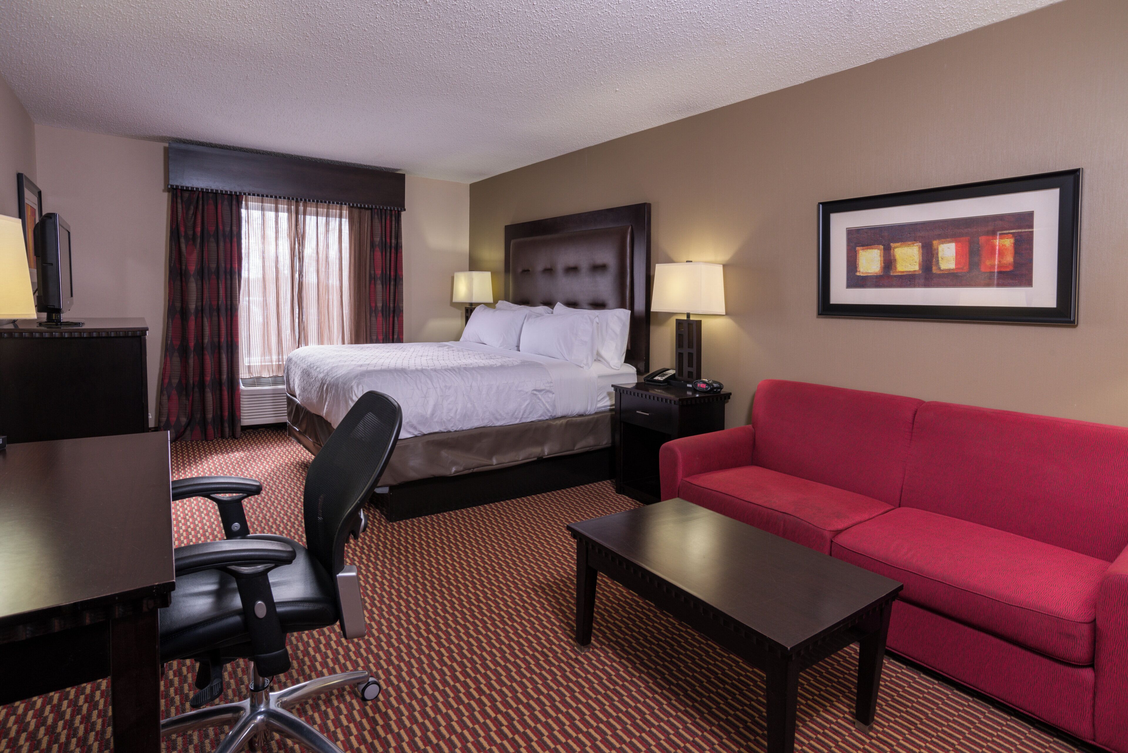 A hotel room with a bed, a red sofa, a desk with a chair, a TV, and a framed picture on the wall.