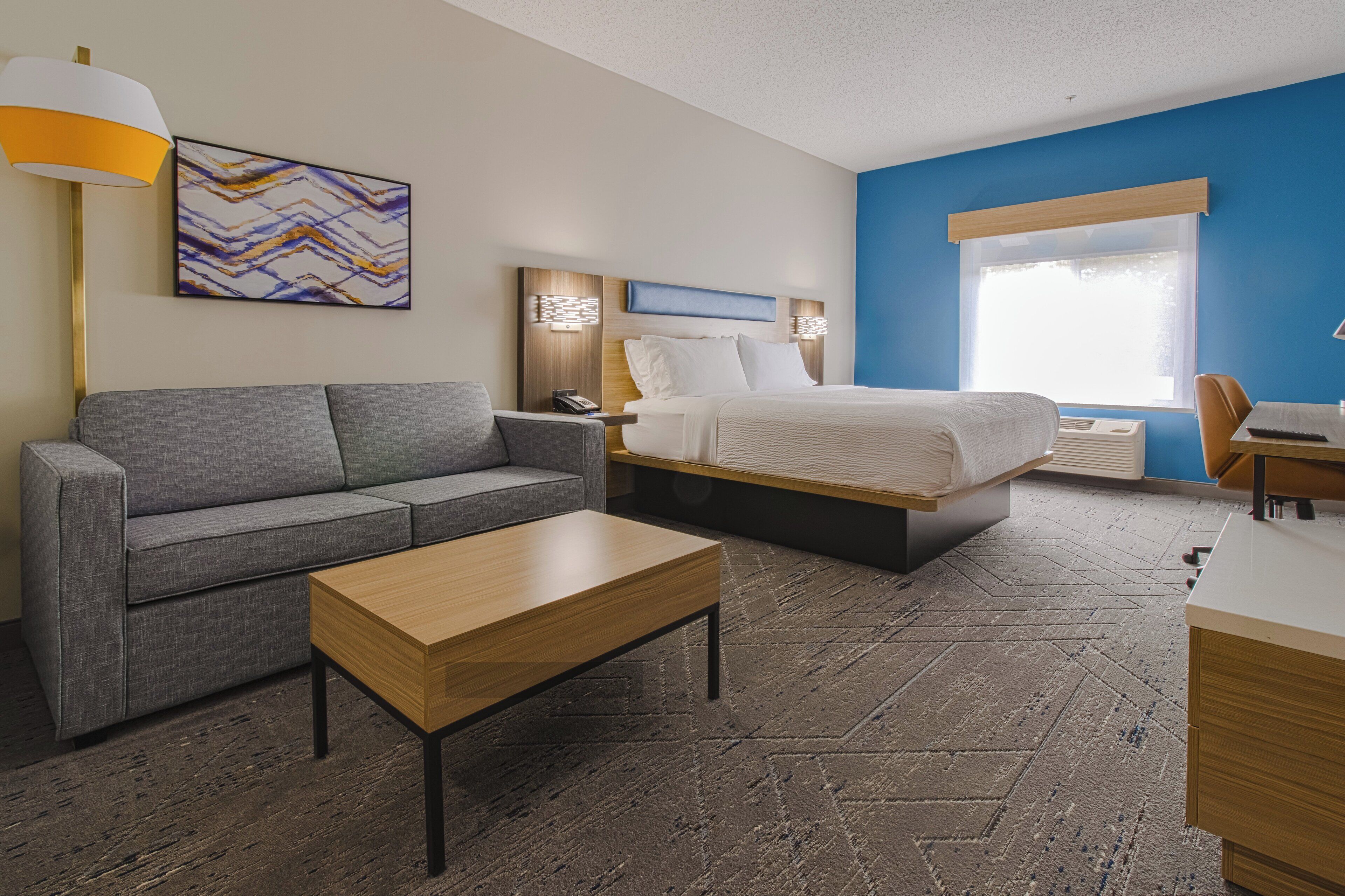 Una habitación de hotel moderna con un sofá gris, una mesa de centro de madera, una cama con edredón blanco y un escritorio con una silla.