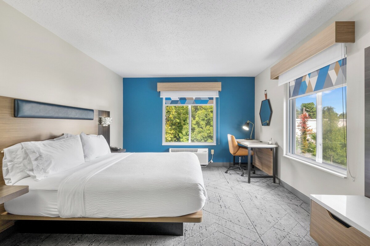 A modern hotel room with a bed, desk, chair, and large windows.