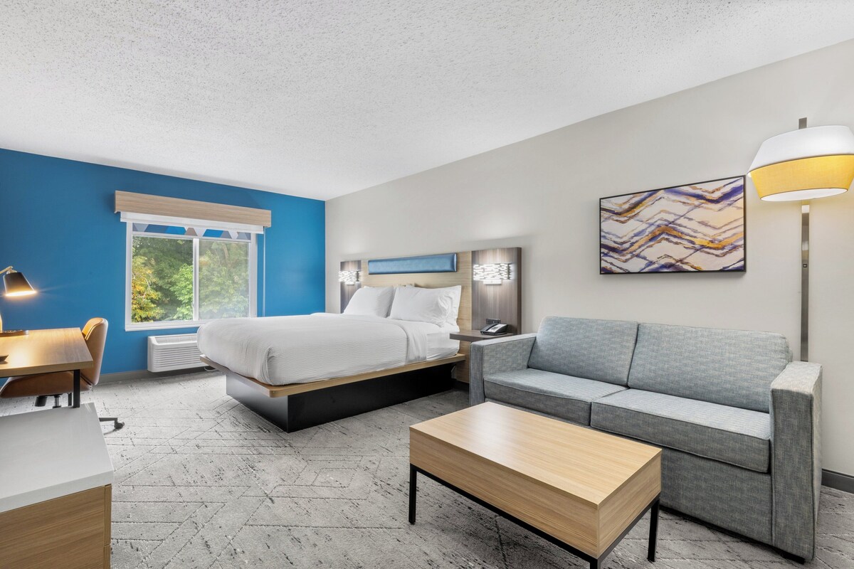 A modern hotel room with a bed, a gray sofa, a wooden coffee table, a desk with a lamp, and a window with a view of greenery.