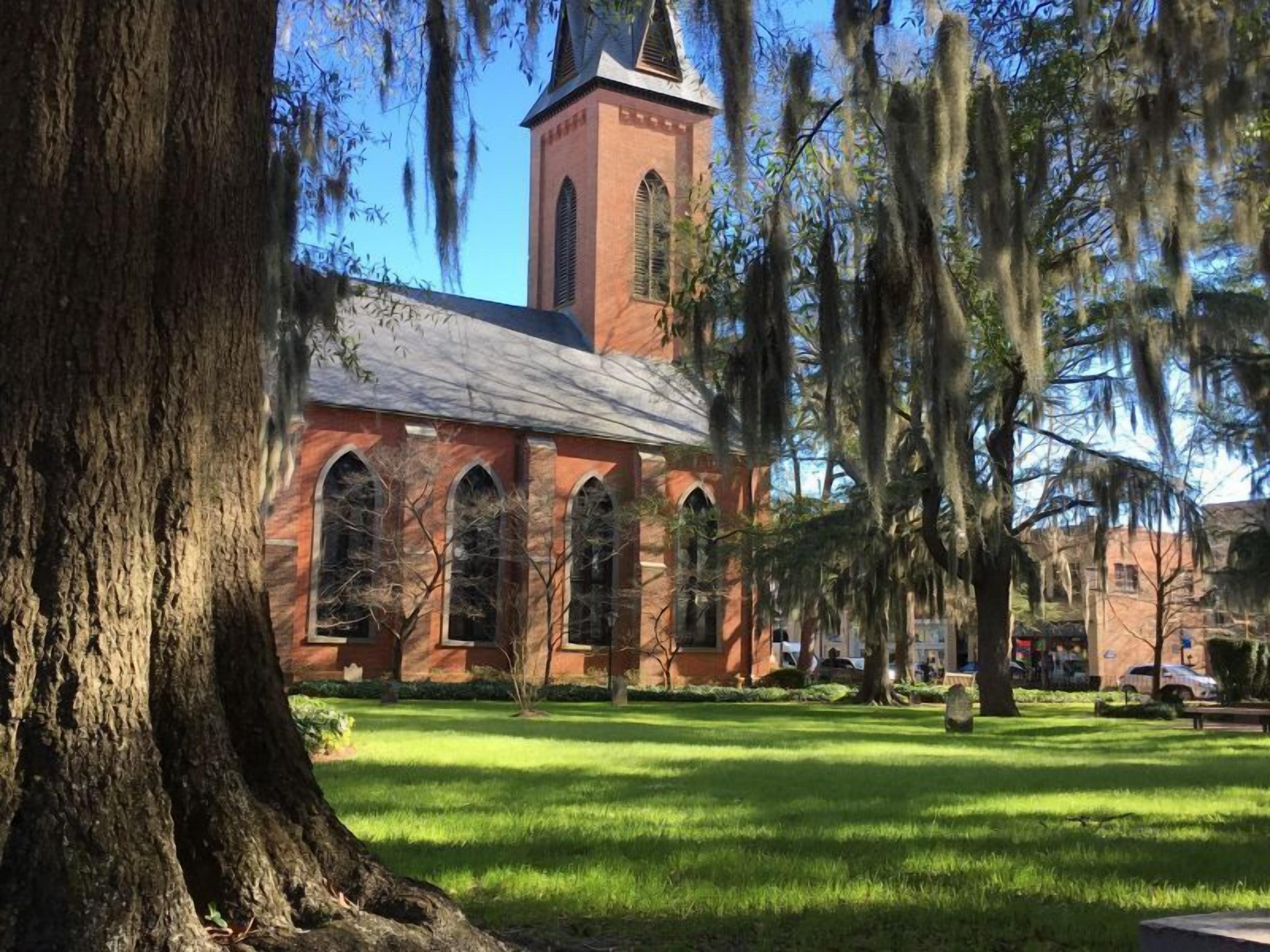 A brick church with arched windows and a steeple, surrounded by trees and grass.