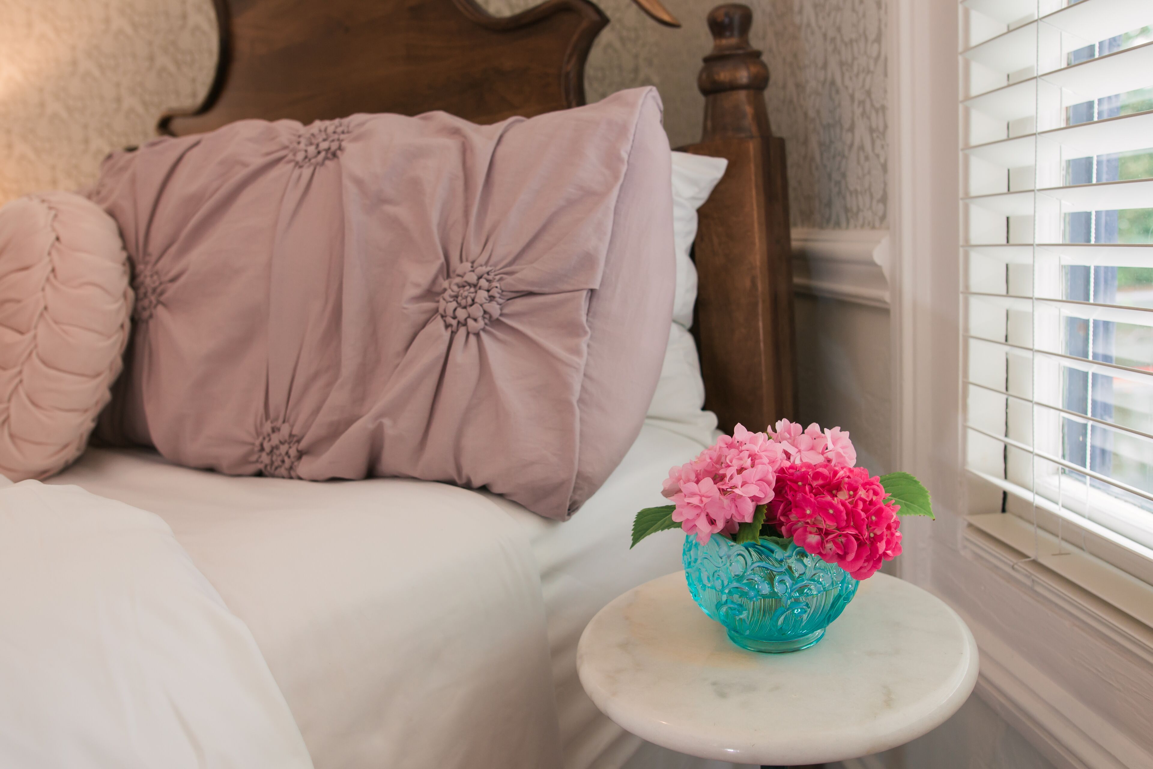 A bedroom with a bed, pillows, a small table with a vase of flowers, and a window with blinds.