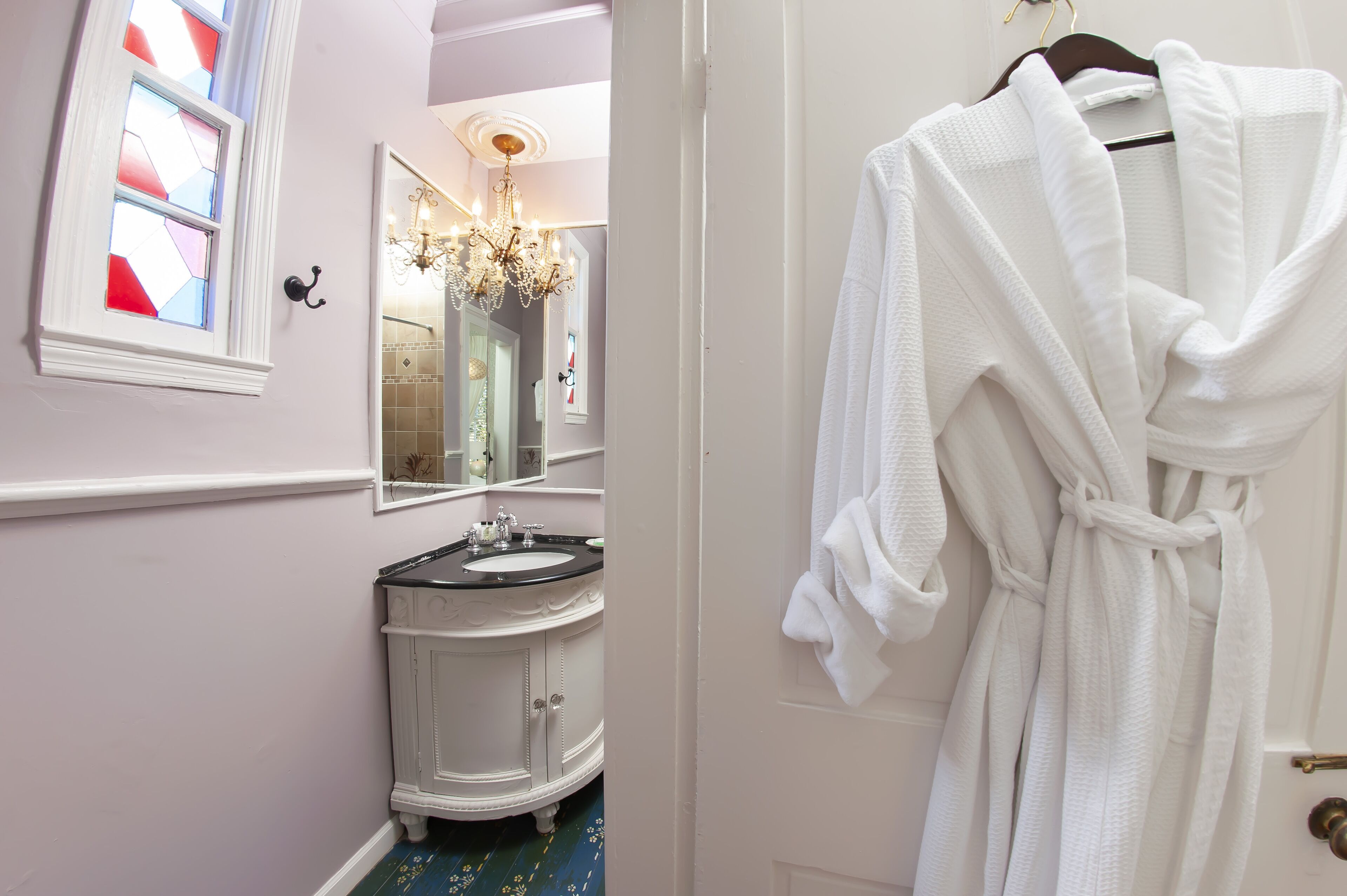 Une salle de bain avec un meuble-lavabo blanc, un miroir, un lustre et une robe blanche accrochée à une porte.
