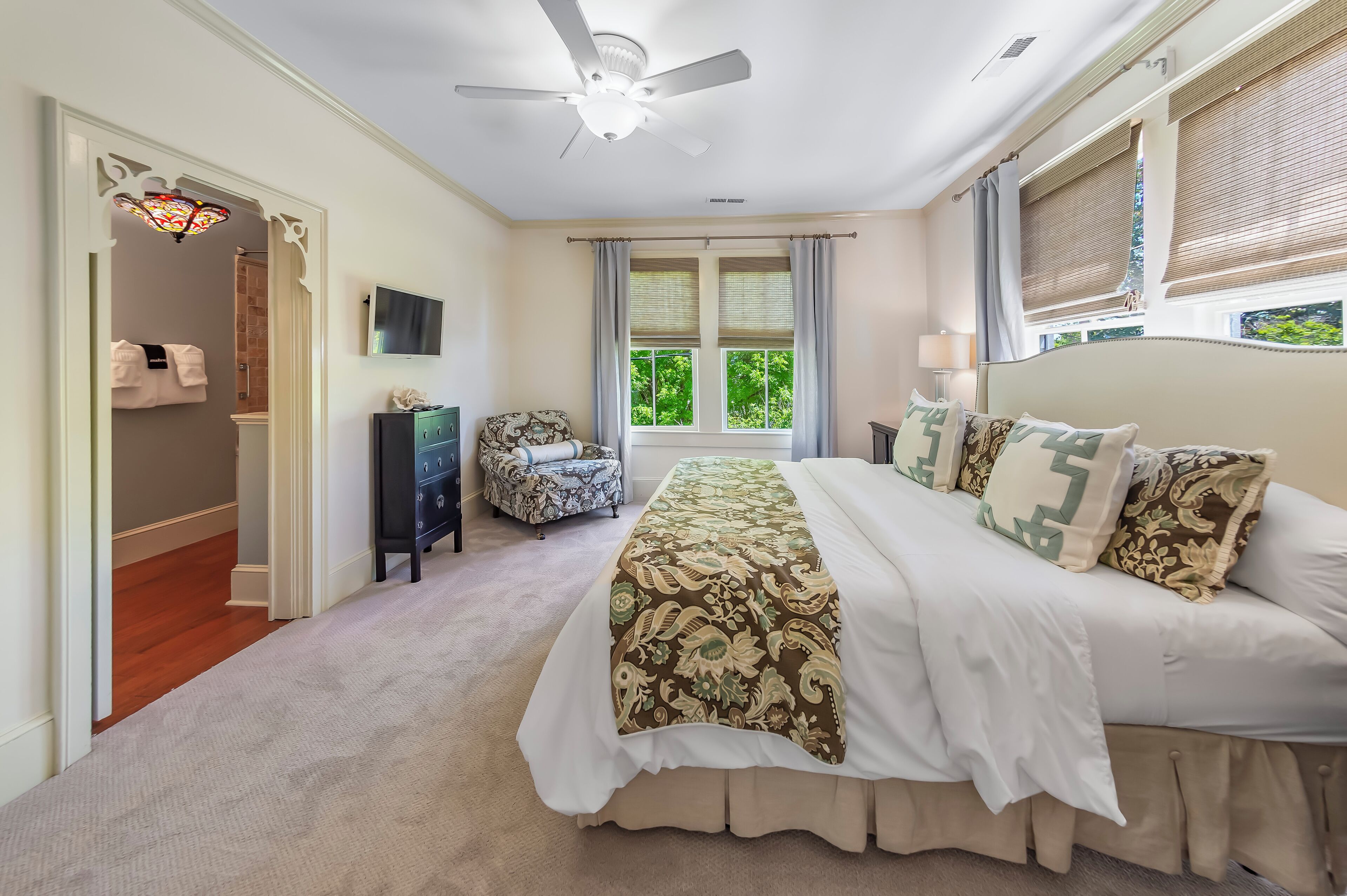 A bedroom with a large bed, a ceiling fan, a TV, a window with curtains, and a door leading to a bathroom.