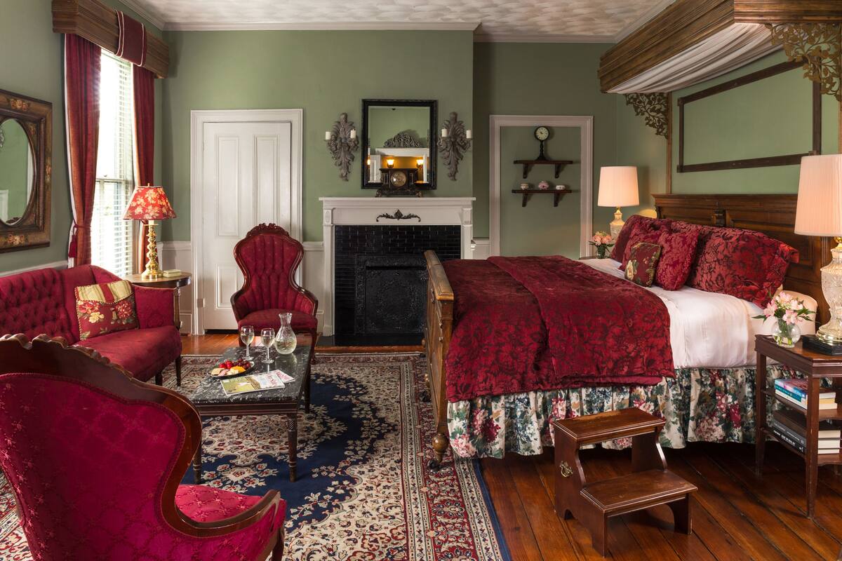 Een klassieke kamer met een open haard, een bed met rode beddengoed, een rode fauteuil en een houten salontafel.