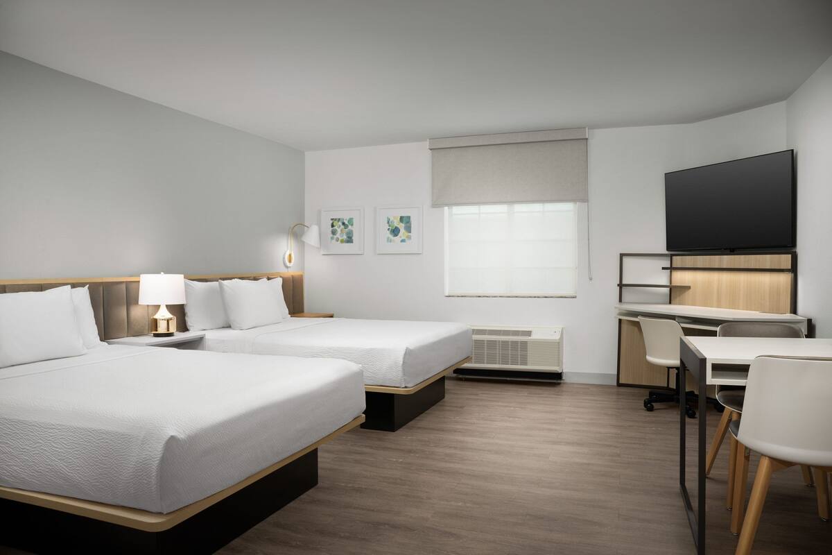 Habitación de hotel con dos camas, un escritorio, una silla, un televisor de pantalla plana y una ventana con persianas.