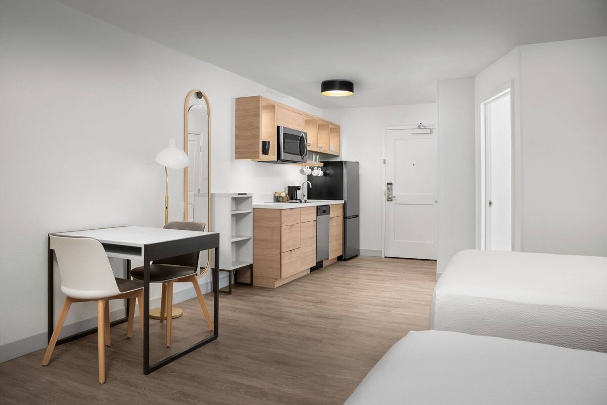 Una habitación de hotel moderna con piso de madera, un escritorio blanco con una silla, una cama, un área de cocina con microondas y refrigerador, y una puerta blanca.