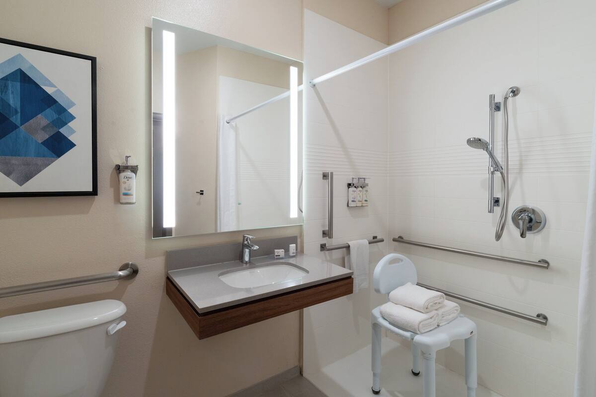 Une salle de bain moderne avec un lavabo, des toilettes et une douche, tous de couleur blanche. On y trouve une barre d’appui, un porte-serviettes et un tableau encadré au mur.