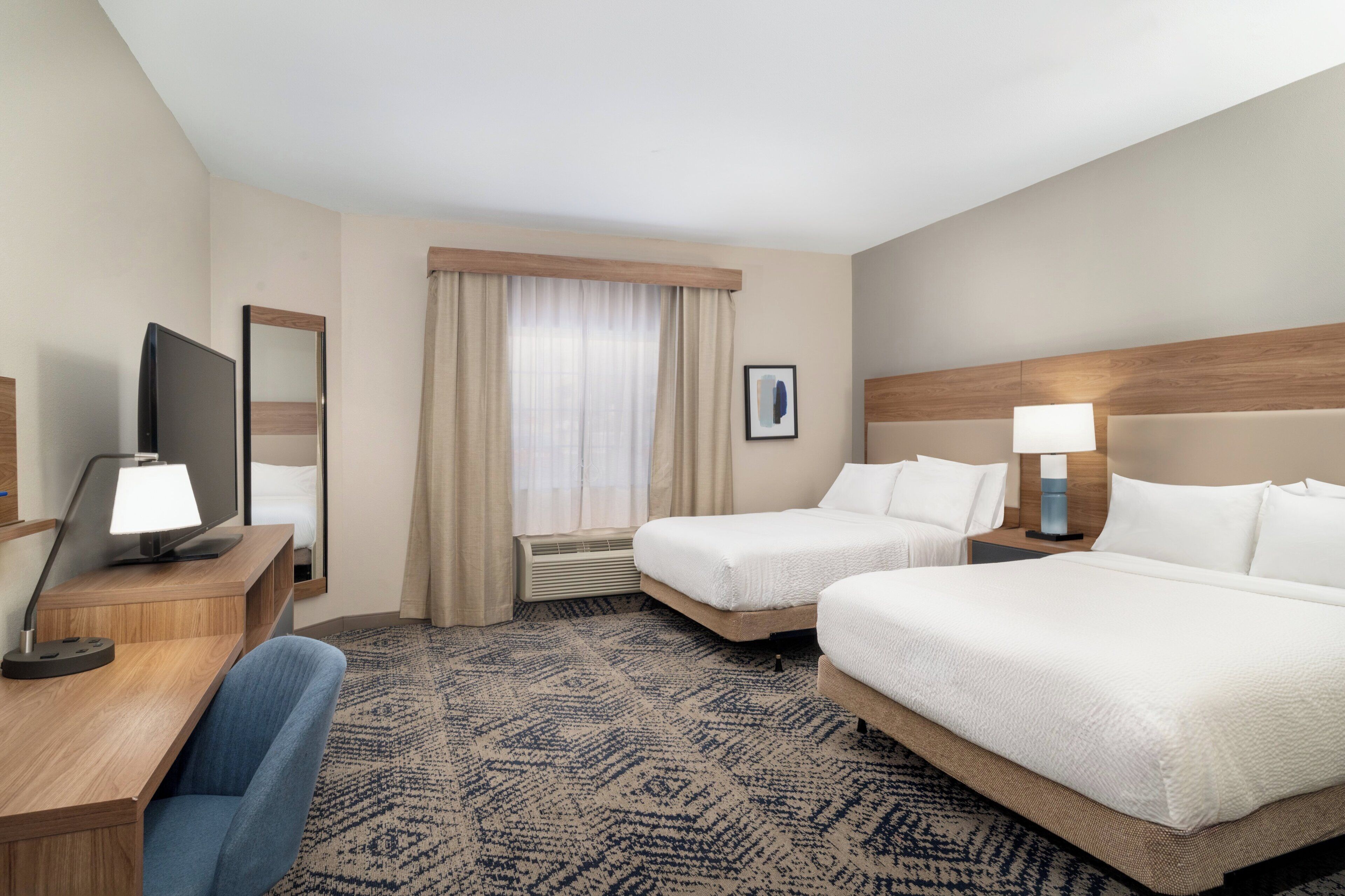 Ein Hotelzimmer mit zwei Betten, einem Schreibtisch mit Stuhl, einem Fernseher, einer Lampe und einem Fenster mit Vorhängen.