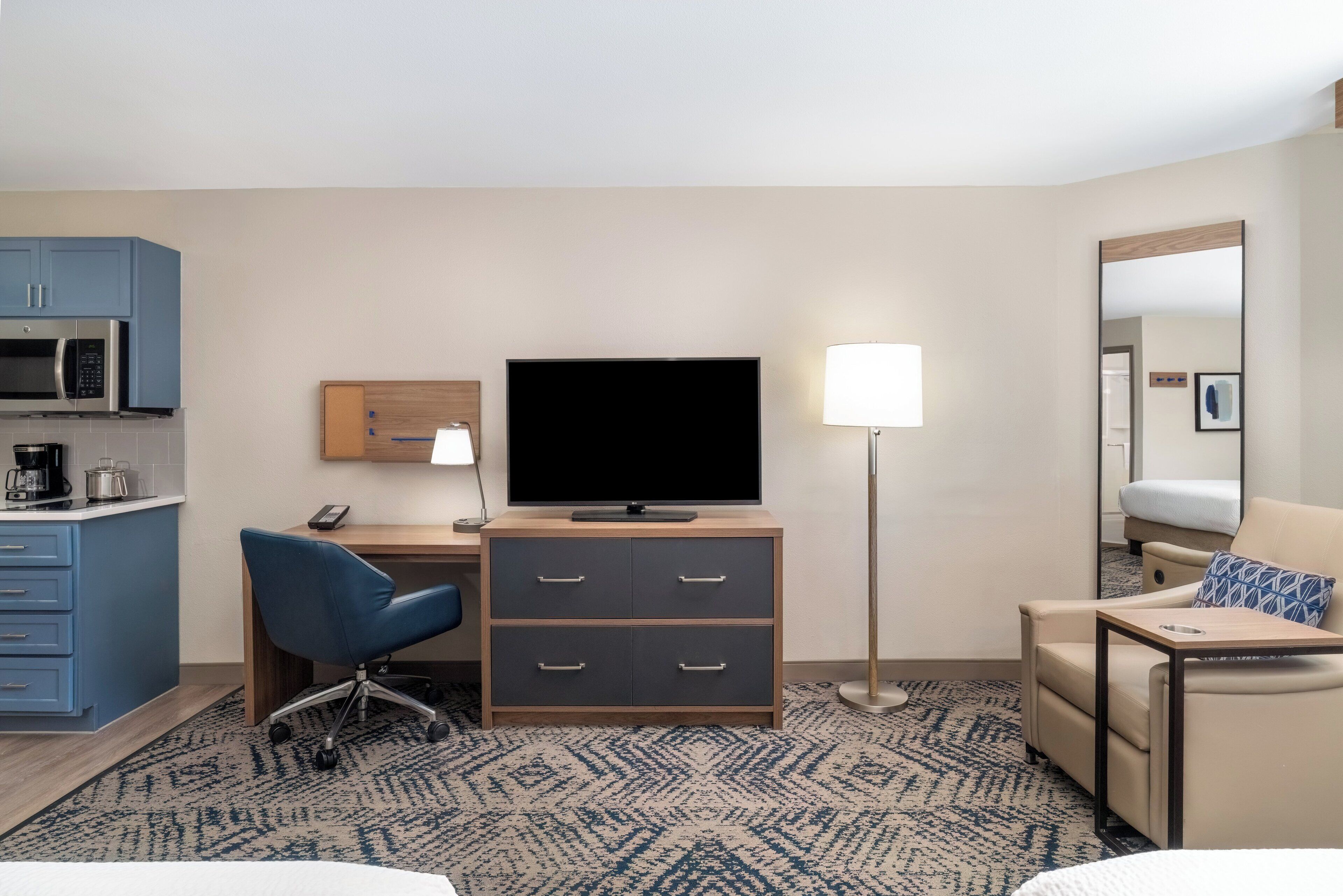 Ein Hotelzimmer mit einer Küchenzeile, einem Schreibtisch mit Stuhl, einem Flachbildfernseher, einer Nachttischlampe, einem Bett und einem Spiegel.