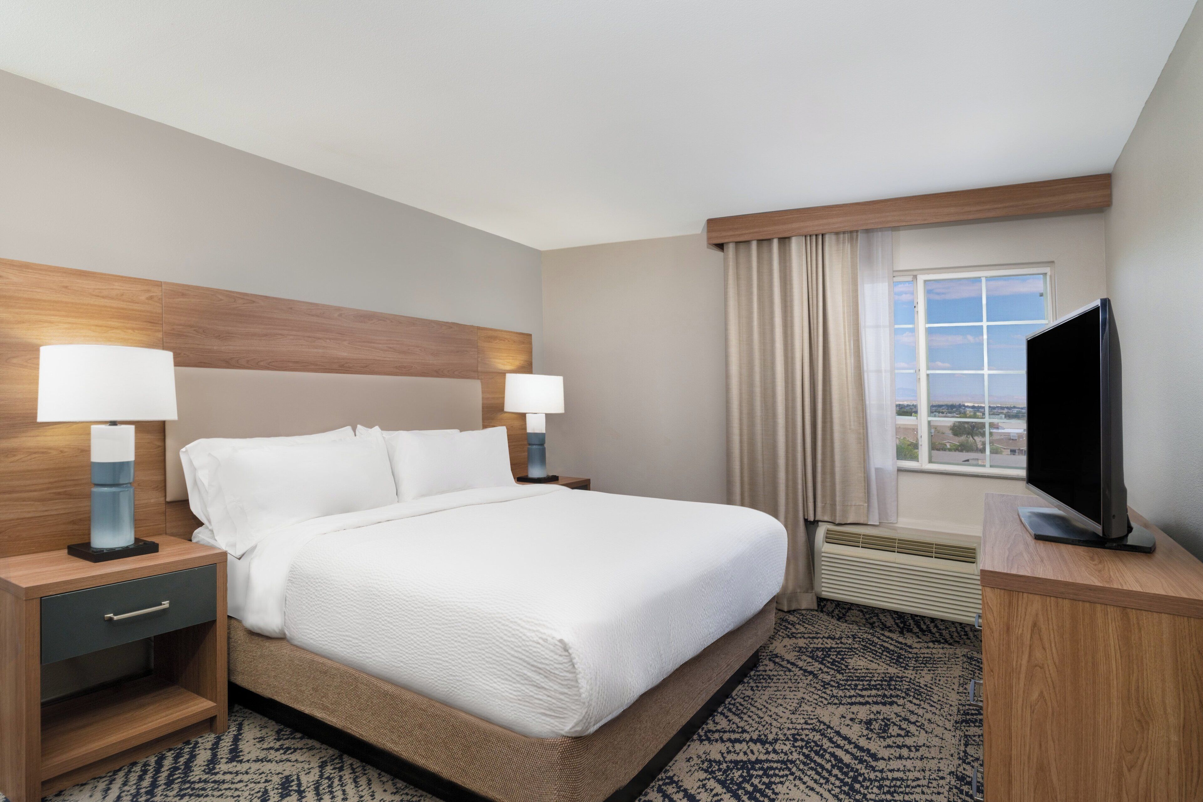 Une chambre d’hôtel avec un grand lit, deux tables de chevet avec des lampes, un téléviseur à écran plat posé sur un meuble en bois, une fenêtre avec des rideaux et une vue sur le paysage urbain.