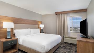 A hotel room with a large bed, two bedside tables with lamps, a flat-screen TV on a wooden stand, a window with curtains, and a view of the cityscape.