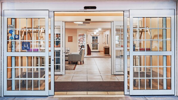 Entrada de hotel con puertas correderas automáticas, zona de recepción y una escalera visible.