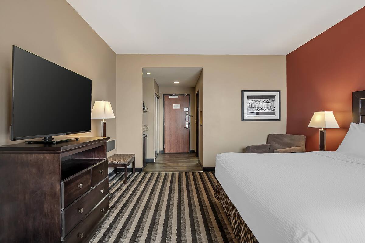 A hotel room with a large bed, a flat-screen TV, a desk with drawers, two lamps, a framed picture on the wall, and a hallway leading to another room.