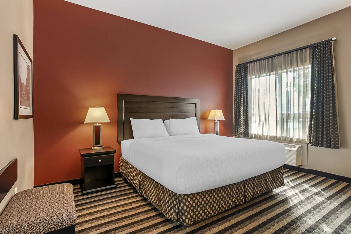 A hotel room with a large bed, two bedside lamps, a small table, a bench, and a window with curtains.