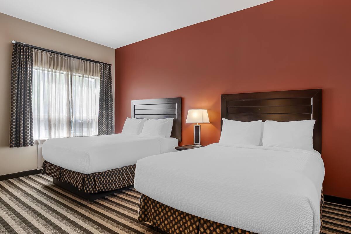 A hotel room with two beds, a striped carpet, a window with curtains, and a nightstand with a lamp.