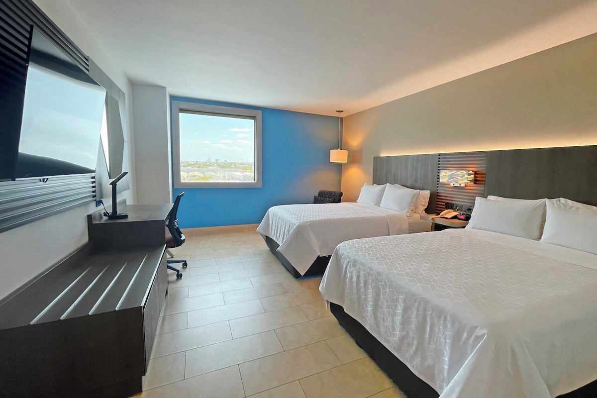 Quarto de hotel com duas camas, uma escrivaninha e uma cadeira. Uma janela oferece vista para a cidade.