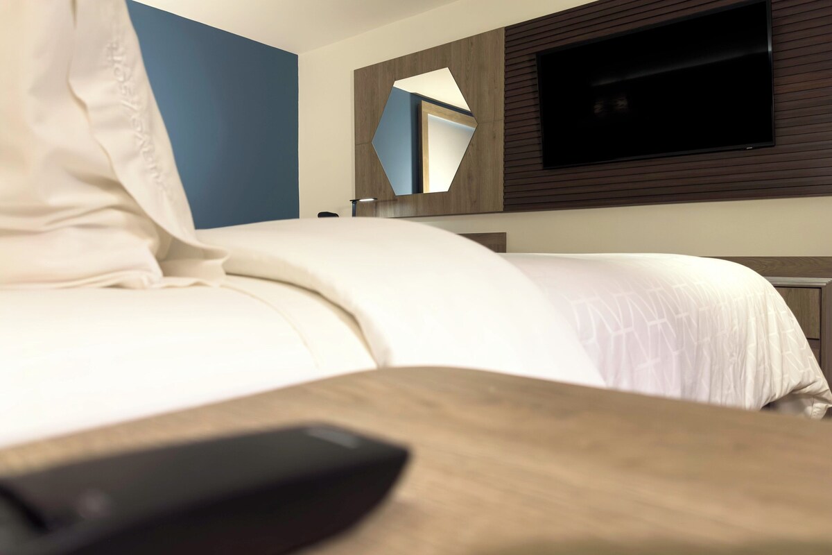 A hotel room with a bed, a TV mounted on the wall, and a wooden nightstand.