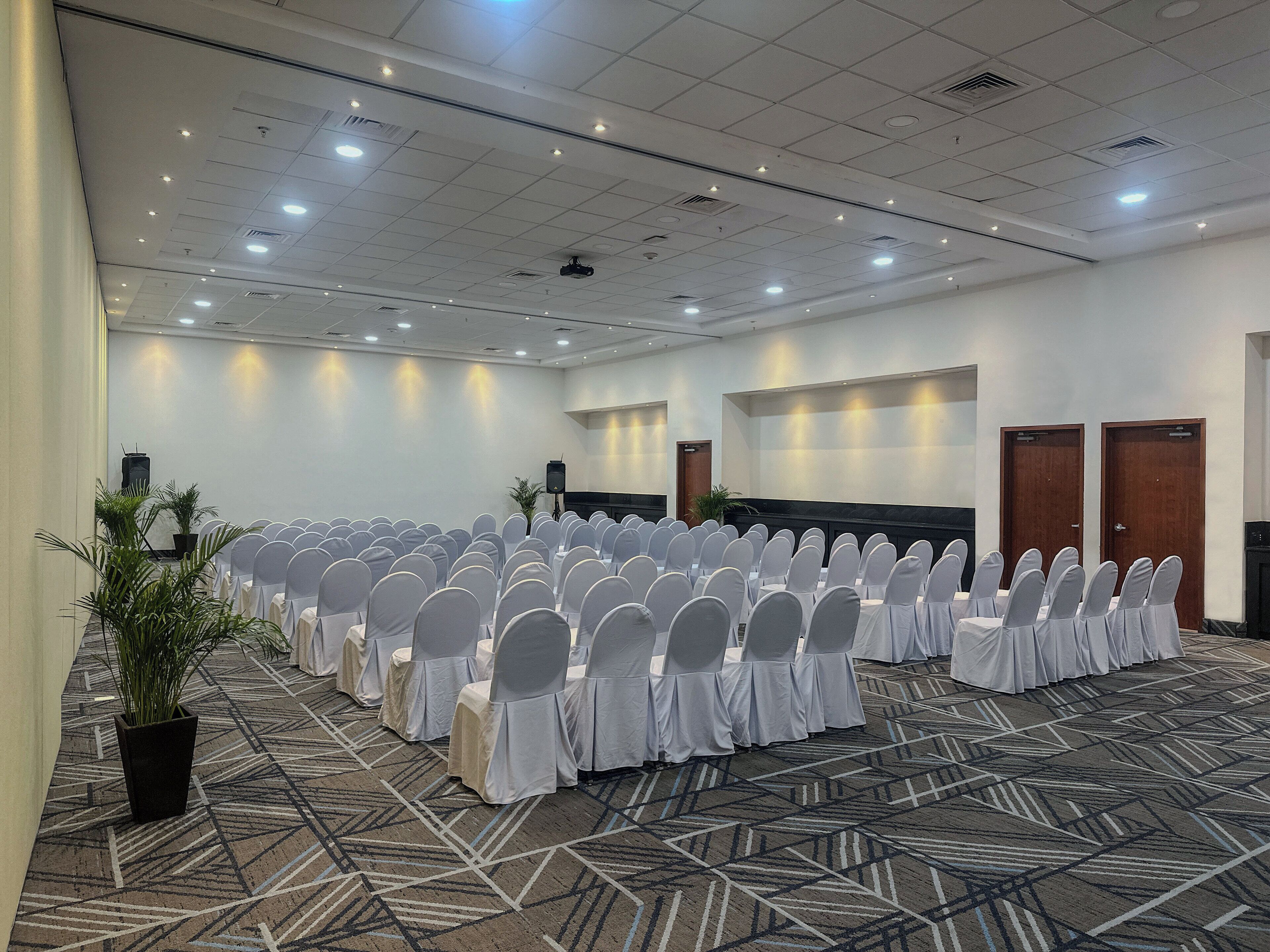 A conference room with rows of white-covered chairs, a stage with a speaker, and a patterned carpet.