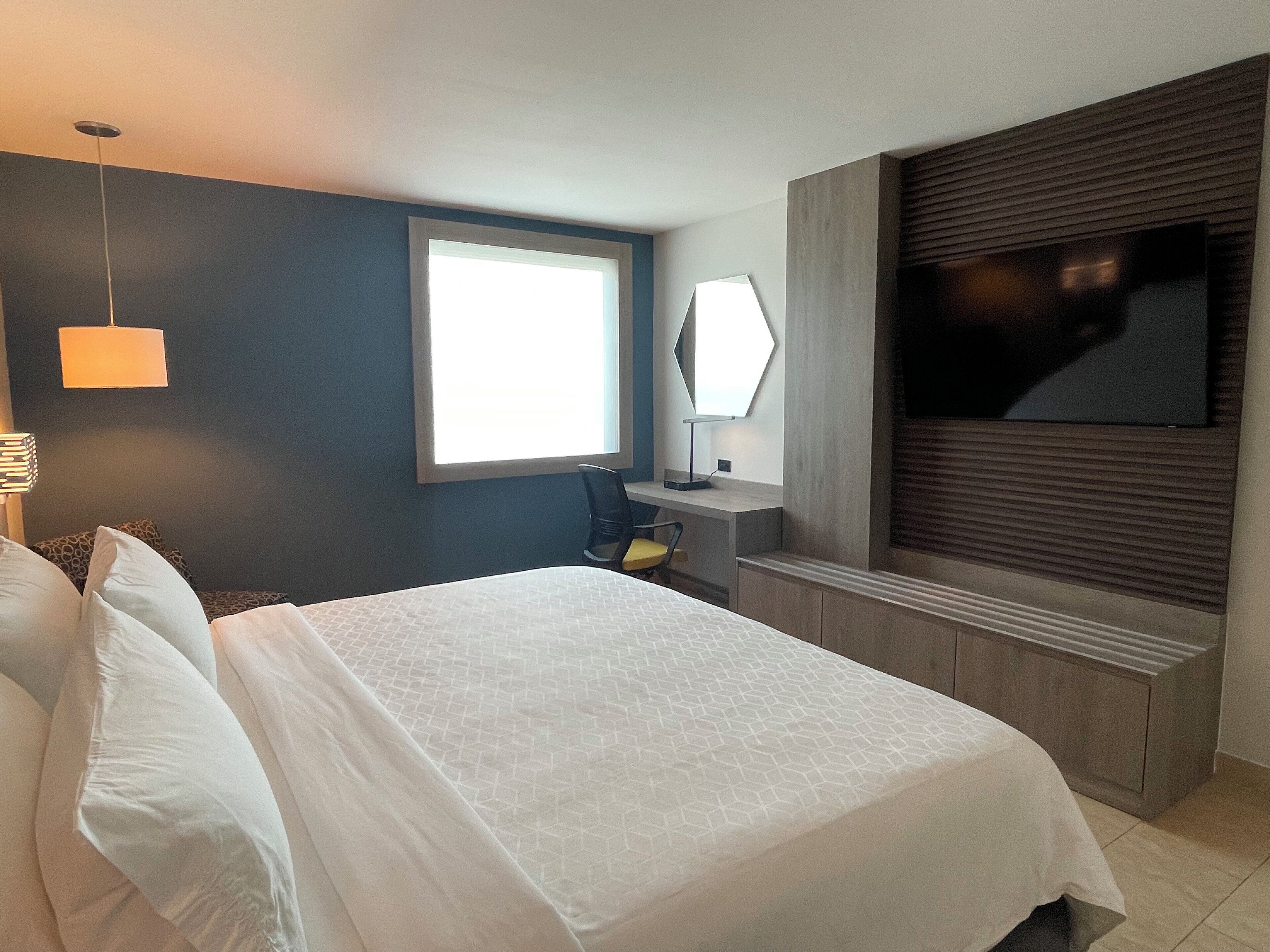 Habitación de hotel moderna con una cama grande, un escritorio, una silla, un televisor de pantalla plana y una ventana que proporciona luz natural.