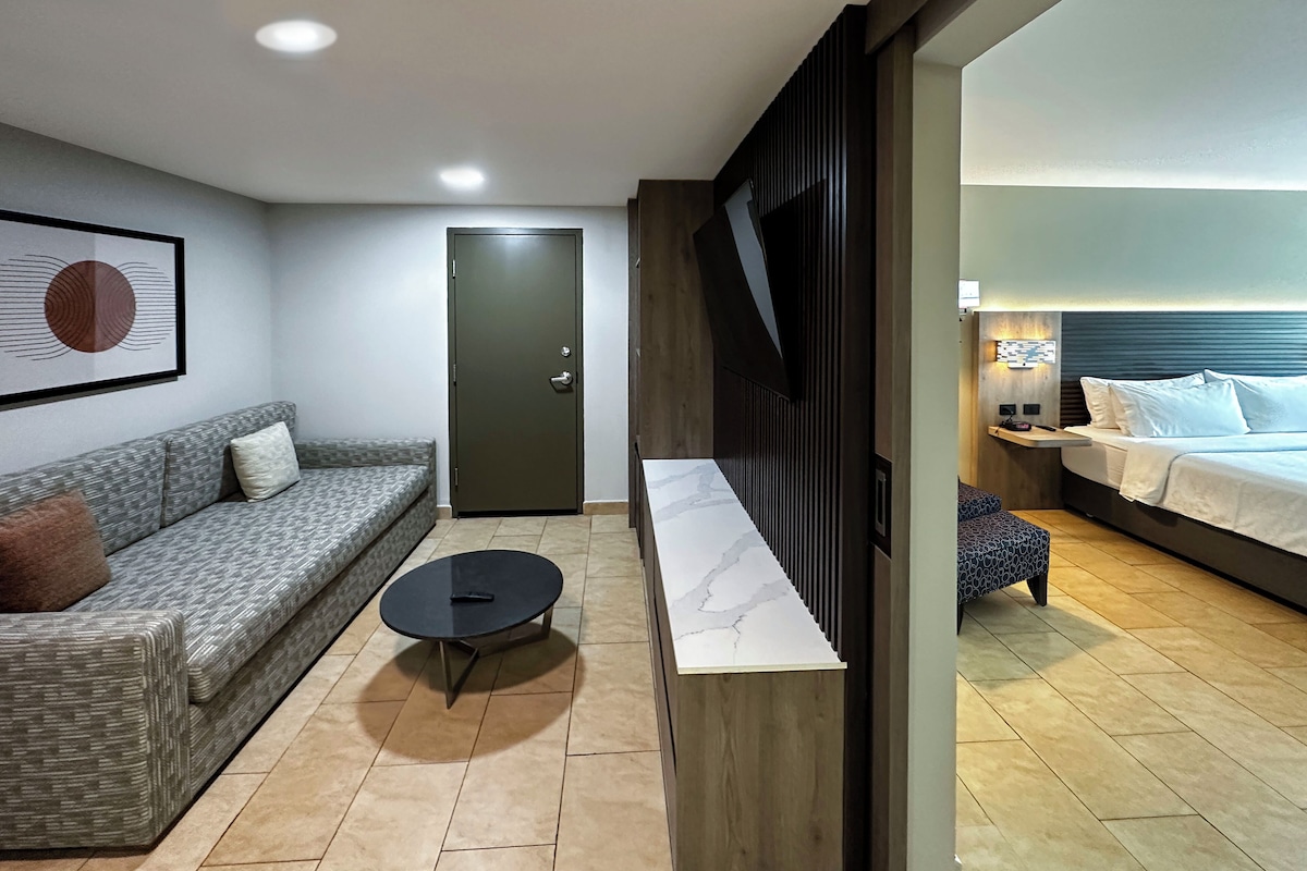 Quarto de hotel moderno com sofá cinza, uma mesa de centro redonda, cama com roupa de cama branca e um divisório de madeira.