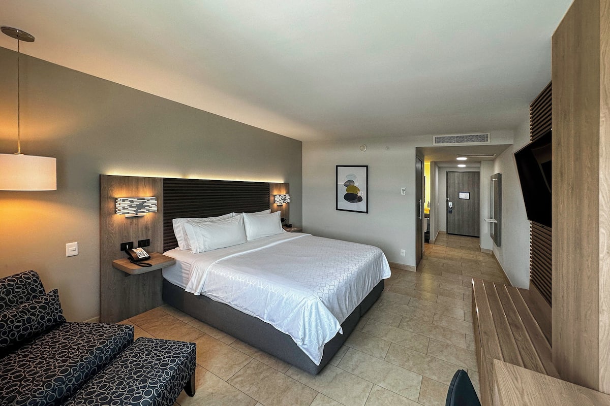 A hotel room with a large bed, bedside tables, a sofa, a TV, and a hallway leading to the bathroom.