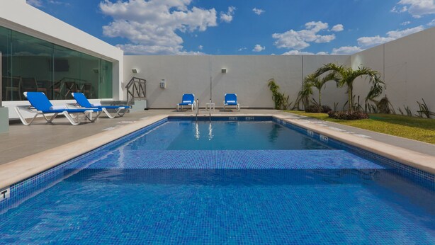 A swimming pool with blue tiles, two blue lounge chairs, and a glass wall on one side. - Holiday Inn Express Mérida by IHG (Mérida)
