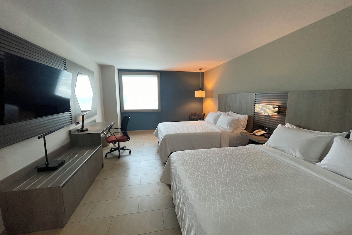 A hotel room with two beds, a desk, a chair, a TV, and a window.
