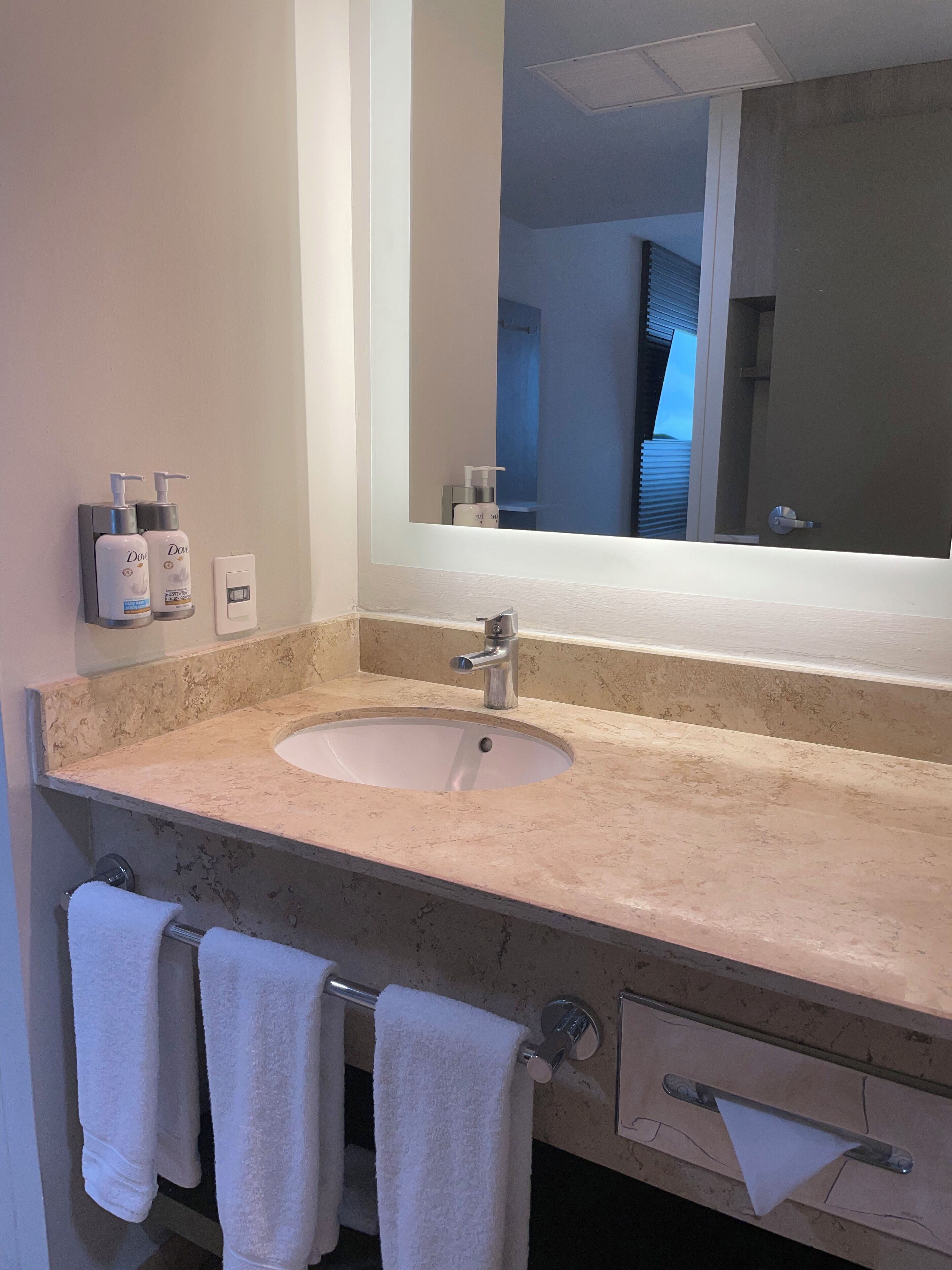 A bathroom with a sink, two hand soap dispensers, a towel rack with three towels, a mirror, and a door with a handle.