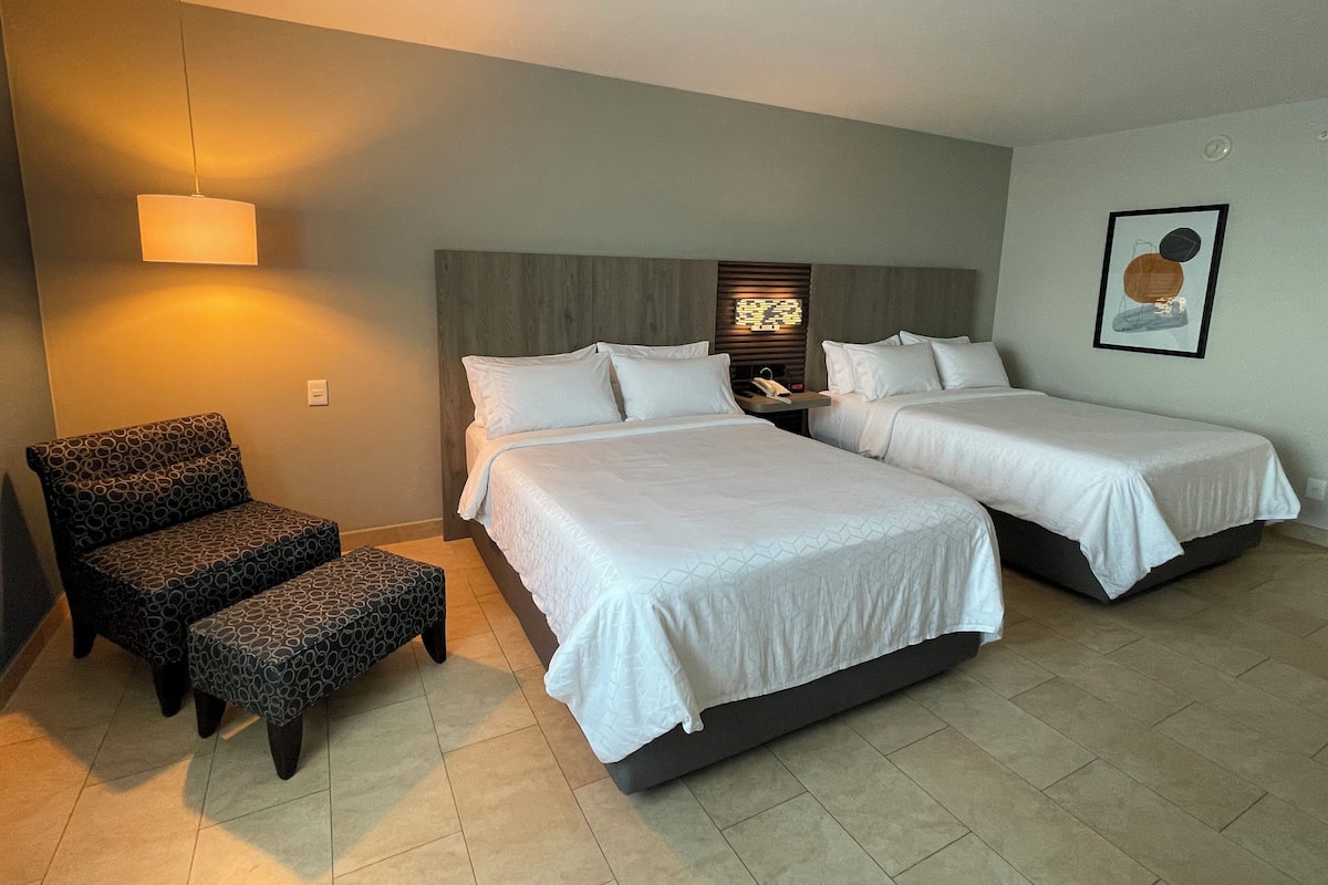 A hotel room with two beds, a patterned armchair, a nightstand, and a wall-mounted light fixture.