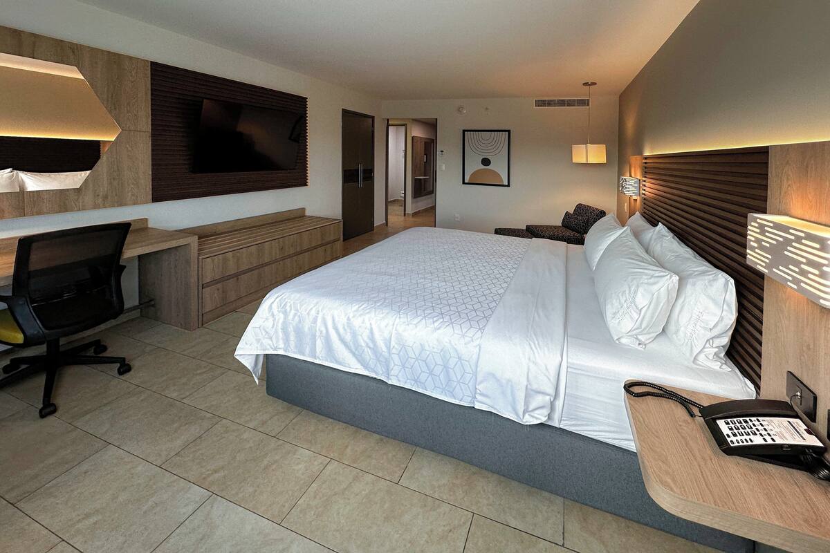 A modern hotel room with a large bed, a desk, a chair, a TV, and a phone on the side table.