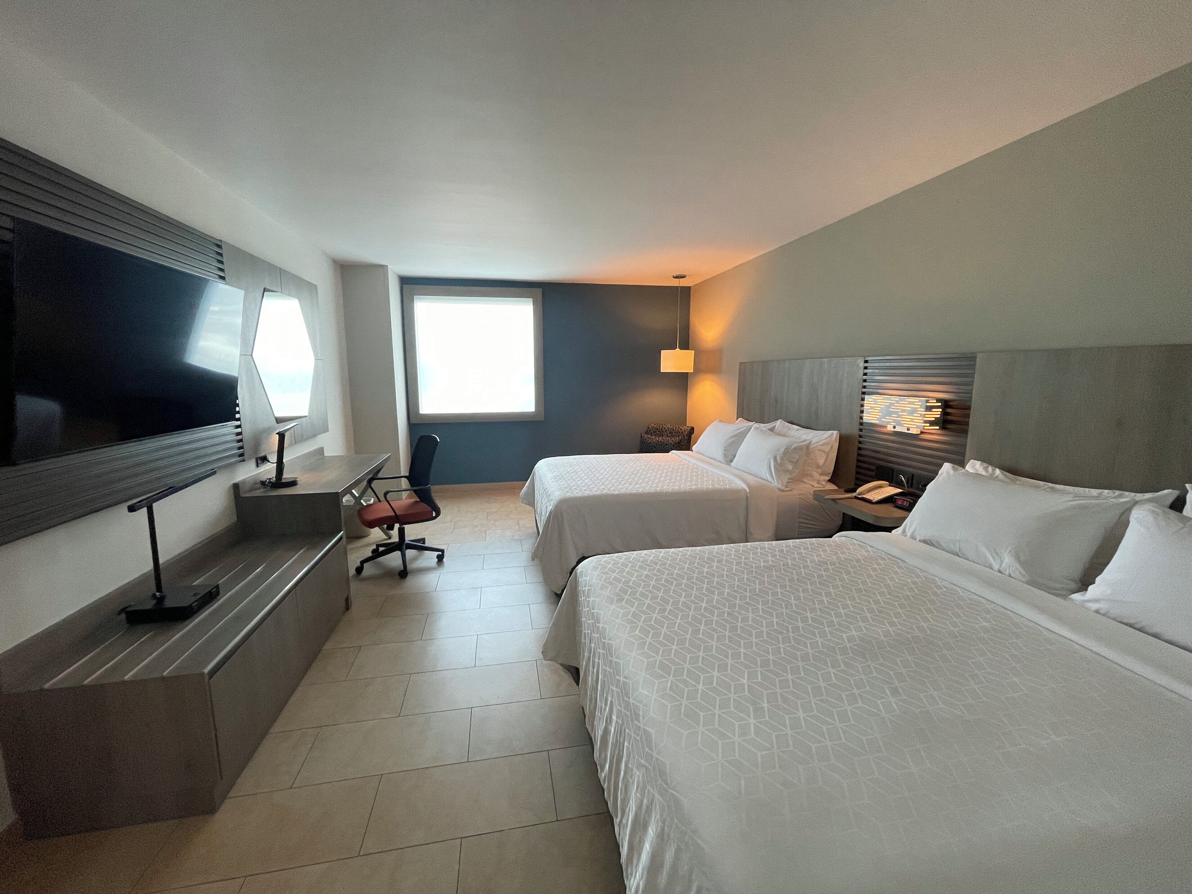 A hotel room with two beds, a desk, a chair, a TV, and a window.