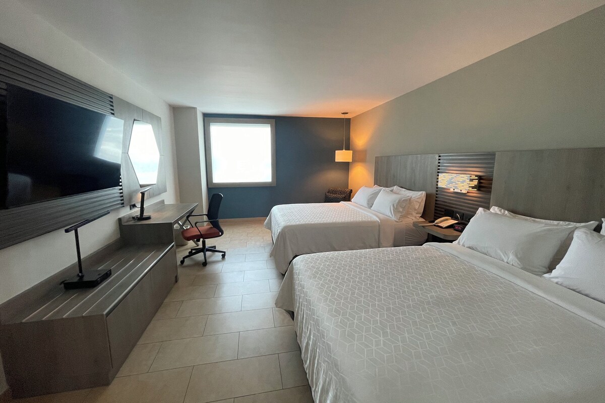 A hotel room with two beds, a desk, a chair, a TV, and a window.