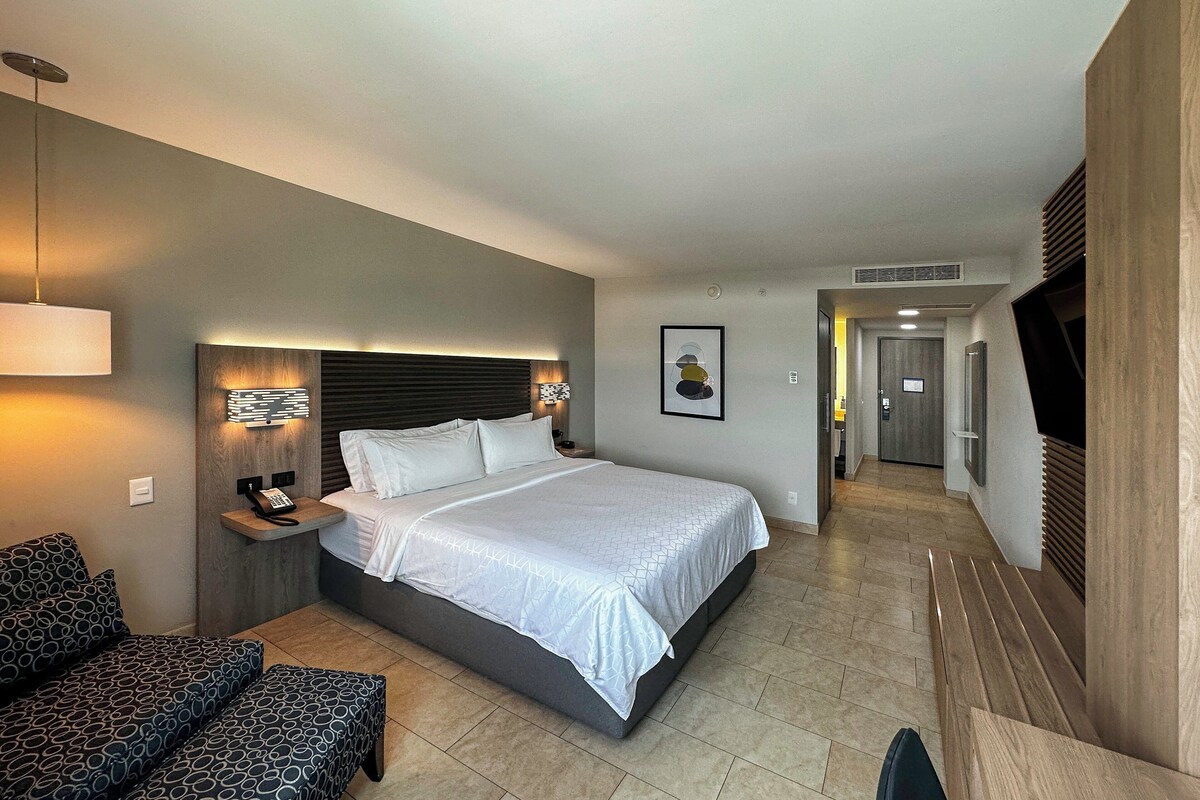 A hotel room with a large bed, bedside tables, a sofa, a TV, and a hallway leading to the bathroom.