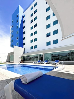 A modern hotel with a swimming pool, blue lounge chairs, and a white building facade.