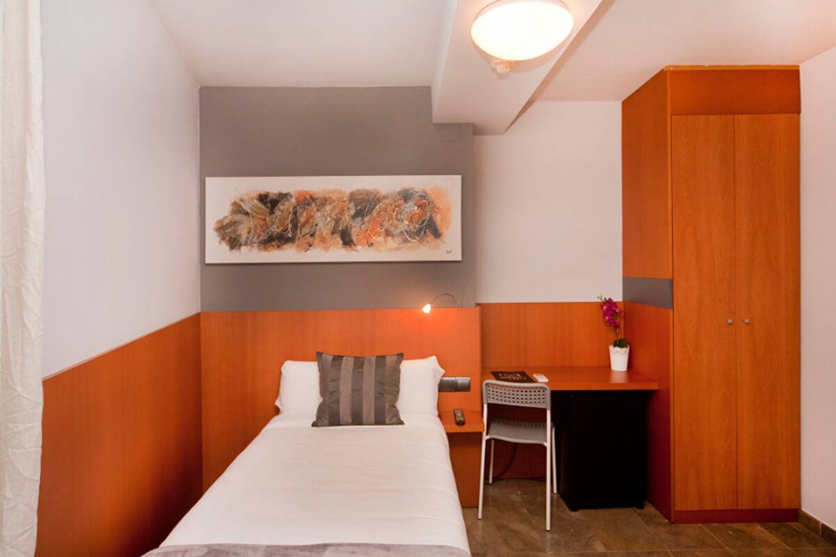A hotel room with a bed, a desk, a chair, a wardrobe, and a painting on the wall.