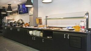 Uma área de buffet com diversas opções de comida, incluindo frutas, doces e bebidas. Há várias estações de serviço com pratos e tigelas, e uma televisão fixada na parede.