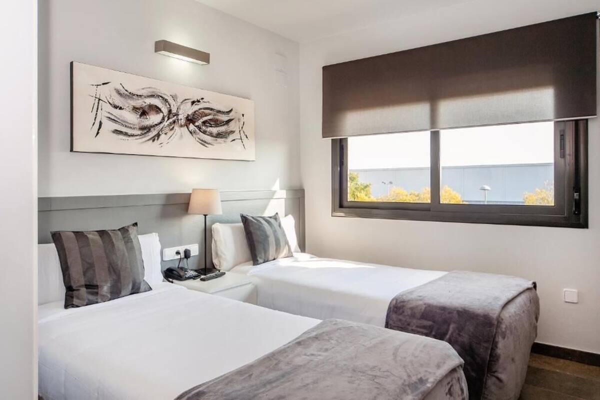 A hotel room with two beds, a large window with a view, a painting on the wall, and a lamp on the nightstand.
