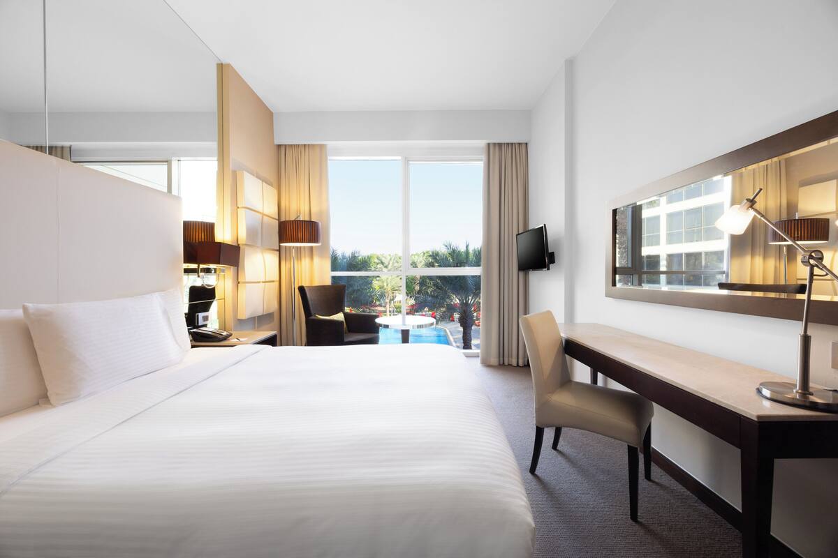 A hotel room with a large bed, a desk, a chair, a TV, and a view of the outdoors.