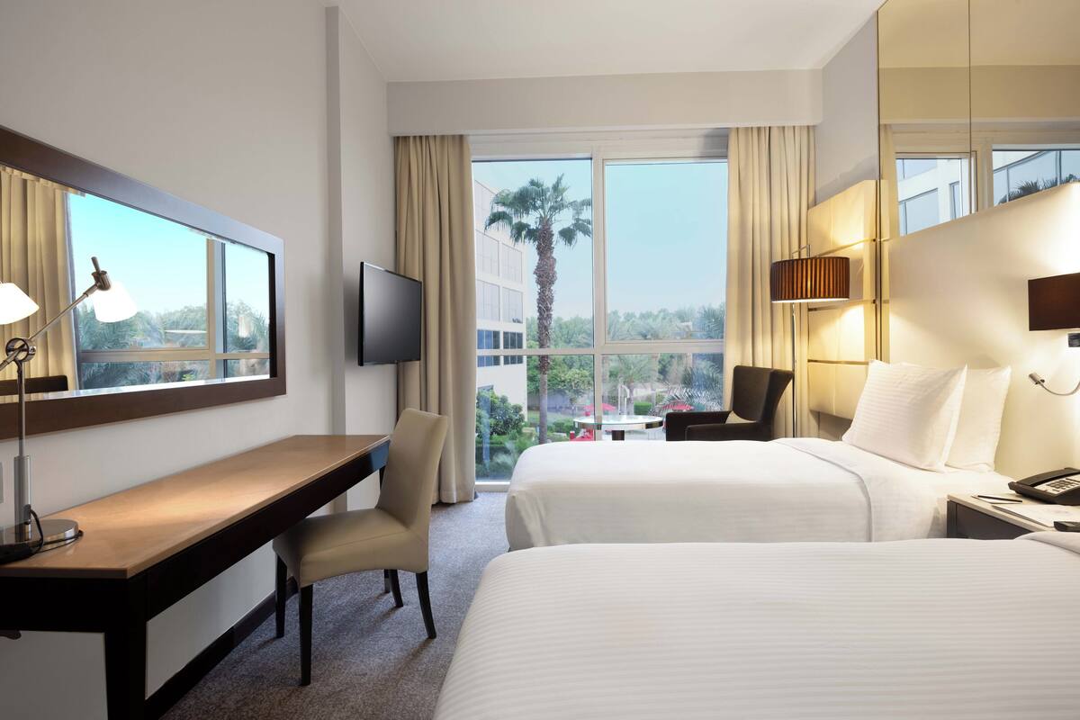A hotel room with two beds, a desk, a chair, a TV, and a large window with a view of palm trees and buildings.
