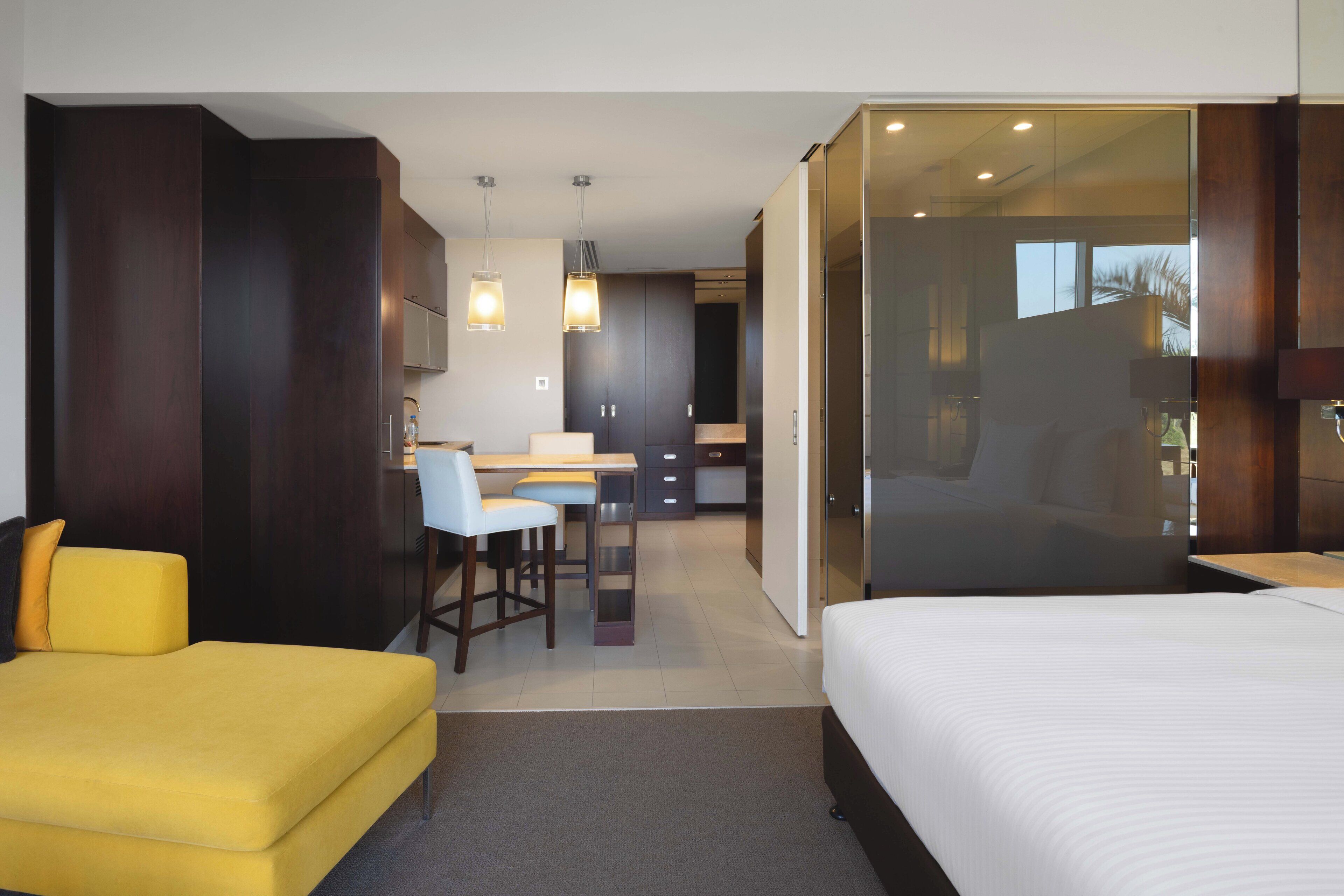 A modern hotel room with a bed, a yellow sofa, a desk with a chair, and a glass partition separating the bedroom from the living area.