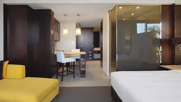 A modern hotel room with a bed, a yellow sofa, a desk with a chair, and a glass partition separating the bedroom from the living area.