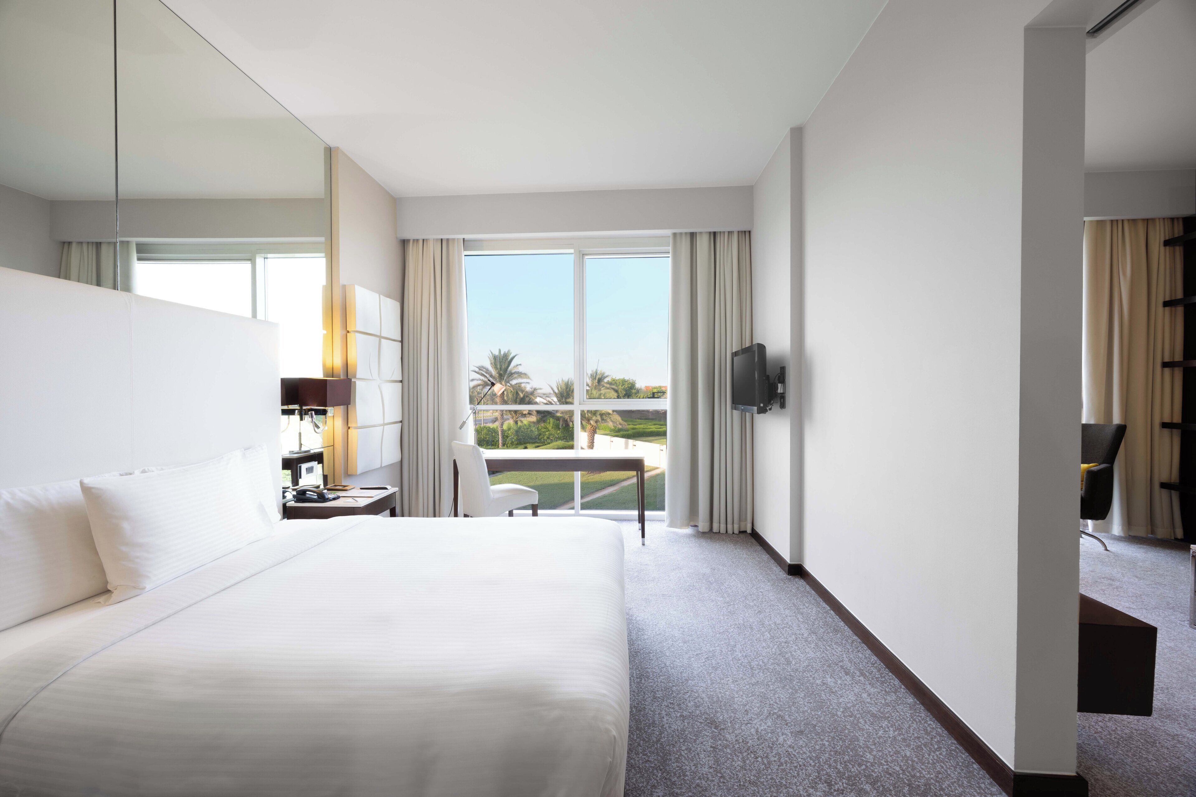 A hotel room with a large bed, a desk with a chair, a television, and a balcony with curtains.