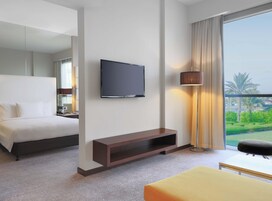 Habitación de hotel con cama, televisor montado en la pared, mesita de noche con lámpara y vista a zonas verdes por la ventana.