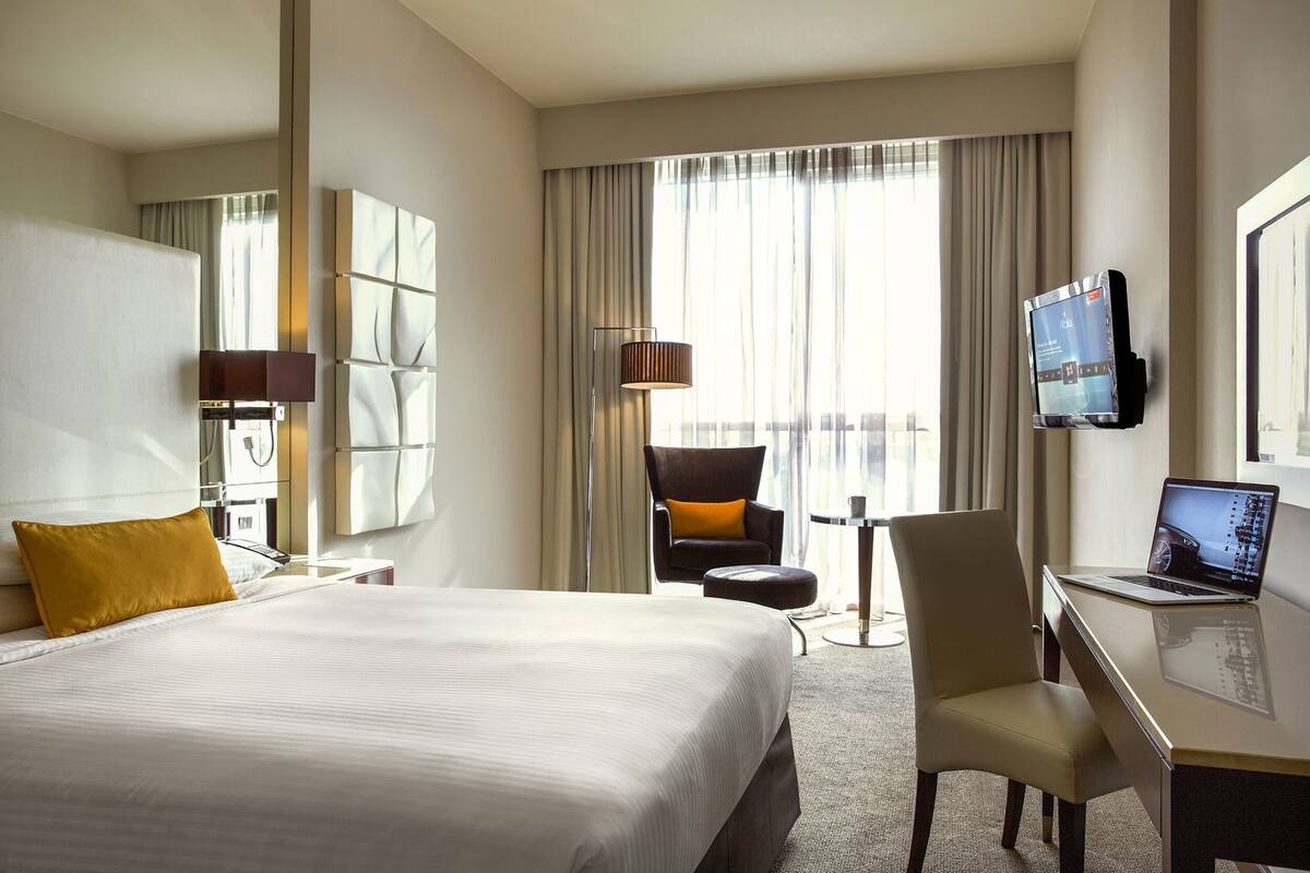 A hotel room with a large bed, a desk with a laptop, a TV, a chair, and a lamp.